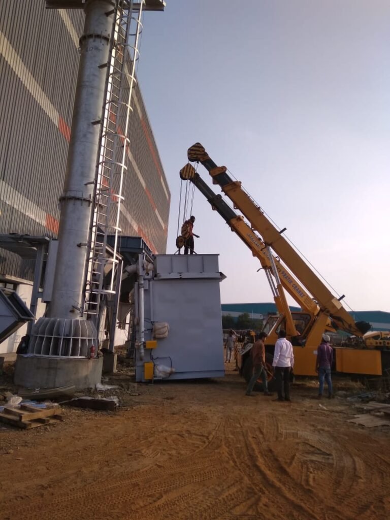 Two cranes erecting a tall steel stack with a ladder beside a plant building