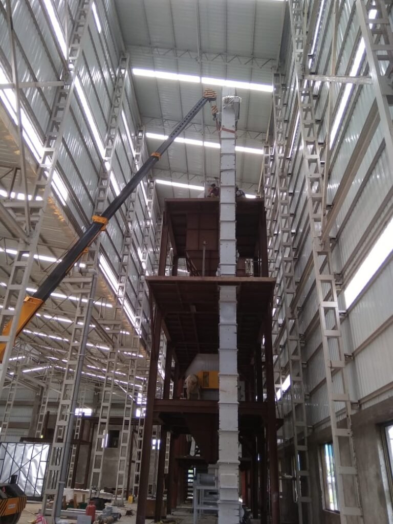 Tall steel tower structure with platforms inside a plant shed, with a crane